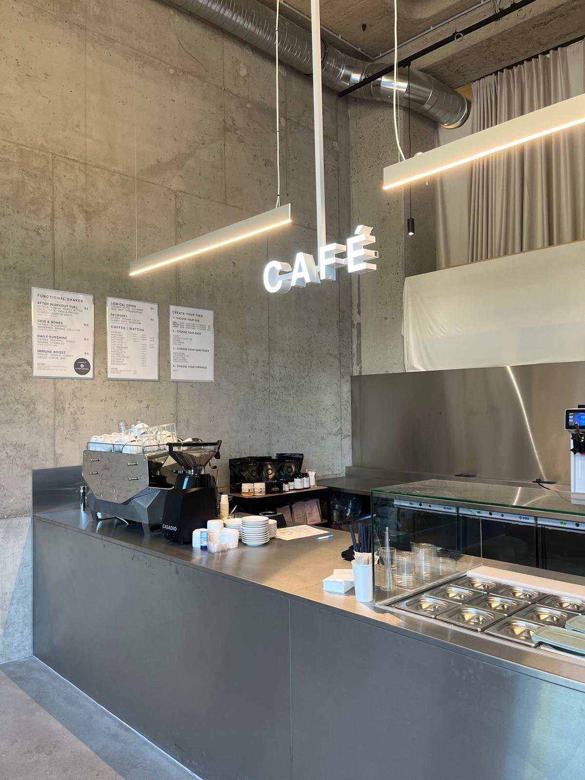 Functional Fuel Café bei ZAYA: Theke mit CAFÉ Leuchtschrift, Espressomaschine und Menükarten an der Betonwand